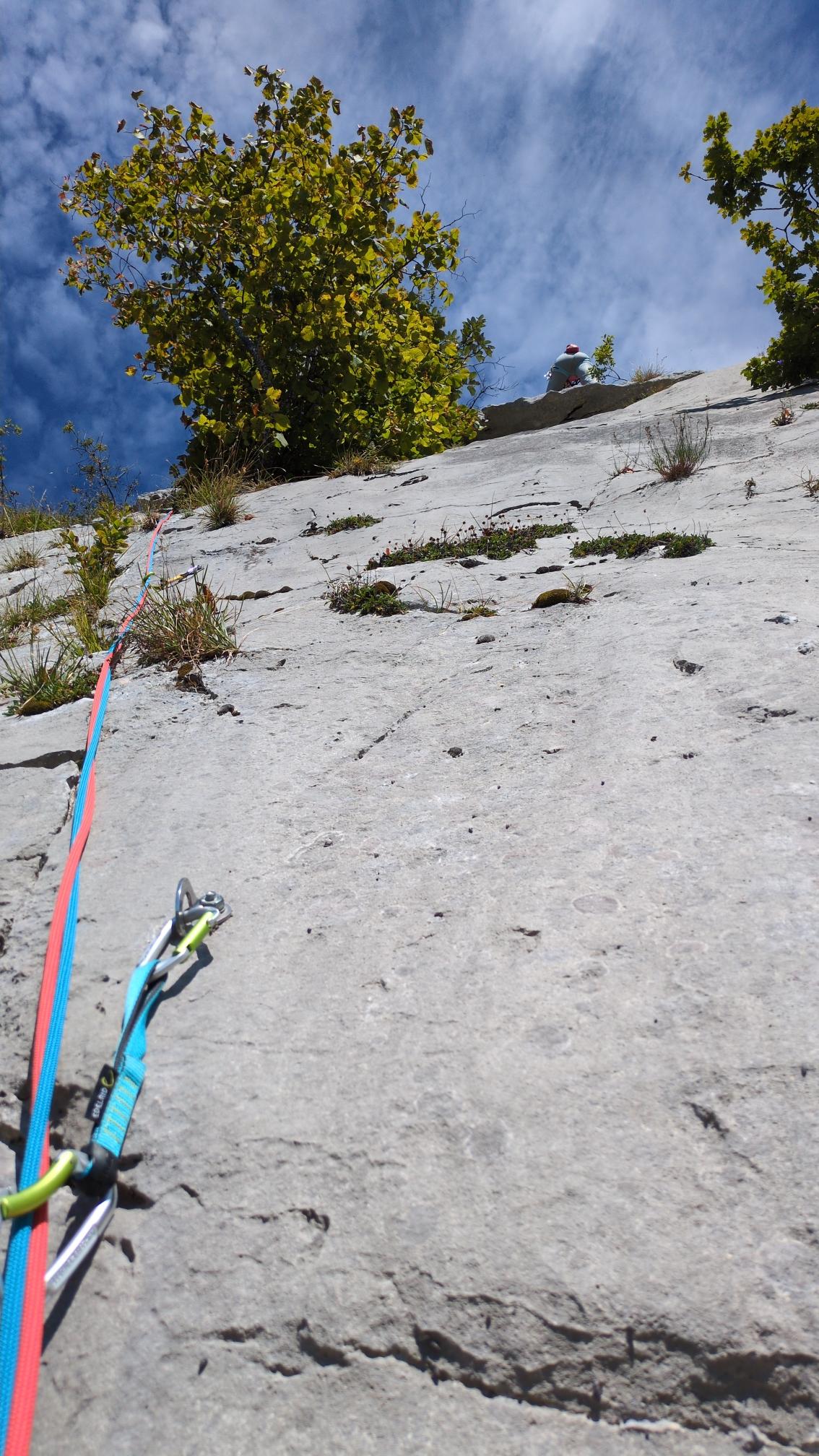A blue and a red rope in an express leading up a grey limestone wall. At the top above a small roof stands my sister in law under a blue sky with thin white clouds.