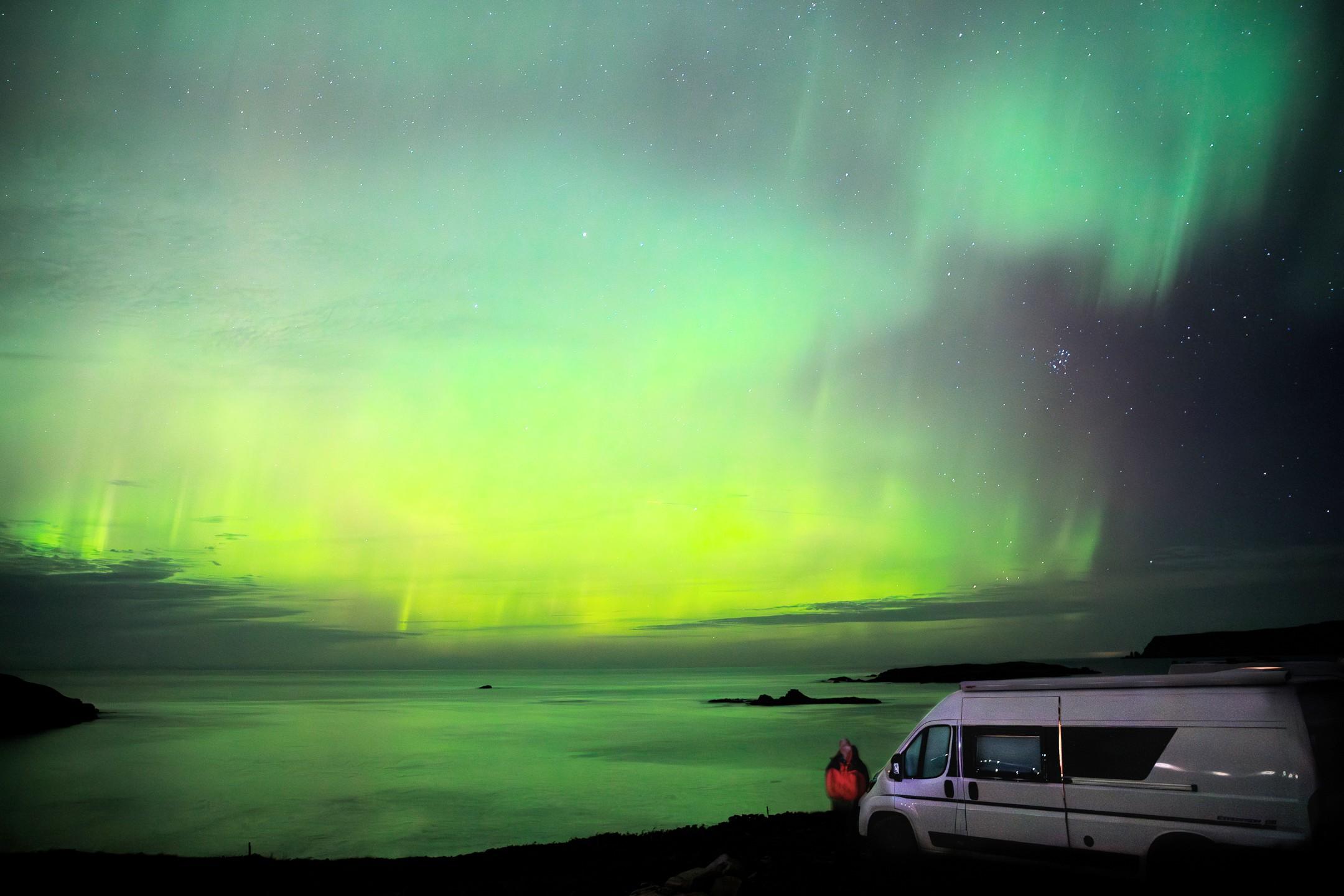 A photograph of Aurora Borealis, the Northern Lights, taken on the north coast of Scotland.
The image is taken out towards the sea. The sky is a big curtain of green, which is reflected in the sea itself. The curtain is ragged on its right hand edge, and the night sky with stars takes up the right edge of the picture.
In the foreground on the right side is a white camper van, pointing leftwards, and standing in front of it is a figure in a red and black jacket. This is me.