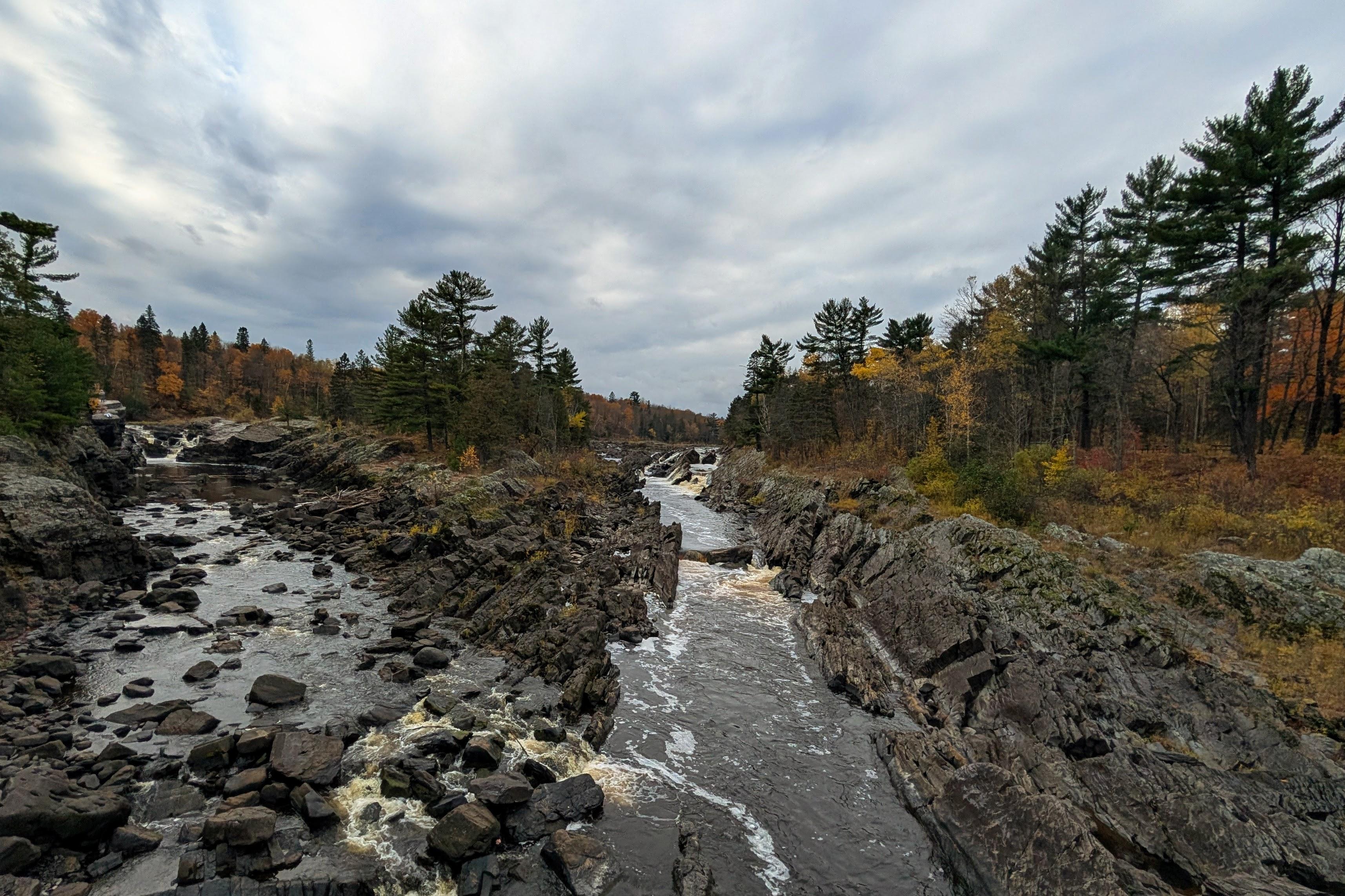 A quick look upstream from the bridge mid-way across. The river has cut two gorges through jagged dark rock, and is coming back together here with just a hint of whitewater over the stones, where normally there are impressive rapids. Tall pines stand green on the far shore, mixed with yellow and orange deciduous trees, not quite as tall.