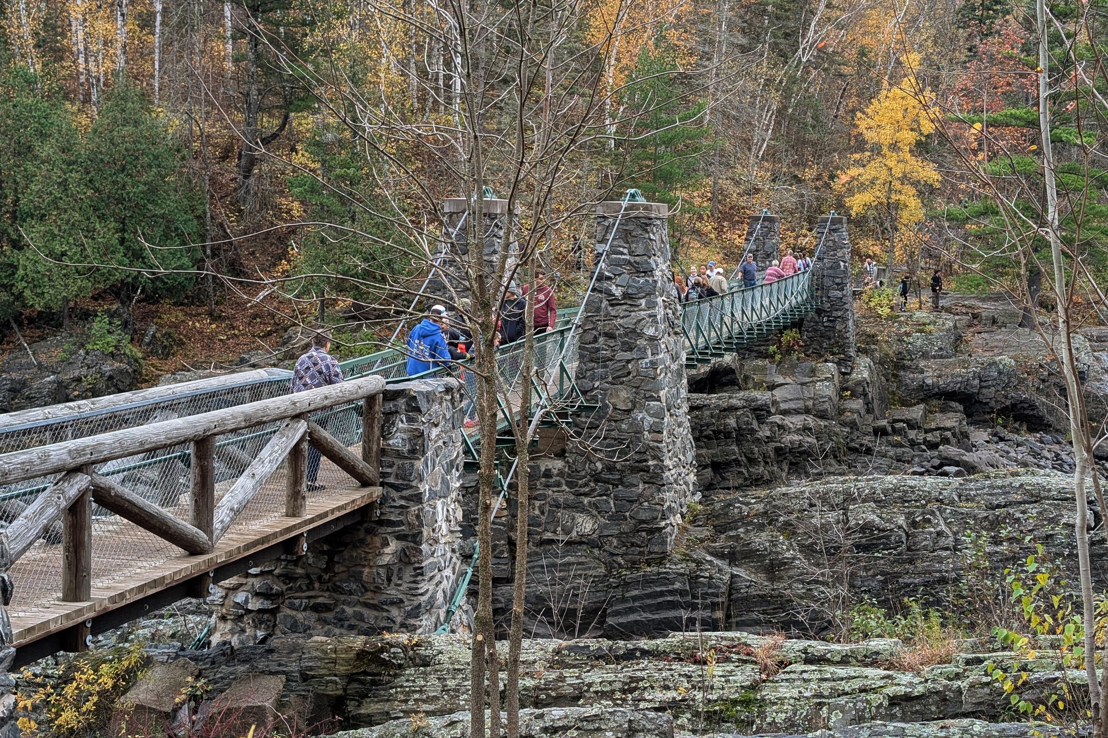 A view of the suspension foot bridge across the St. Louis river gorge, which cuts through jagged dark bedrock. The same dark stone was used to create rock pillar supports for the bridge, which is barely wide enough for people to pass one another going each way. The bridge is full of people across the main span.
