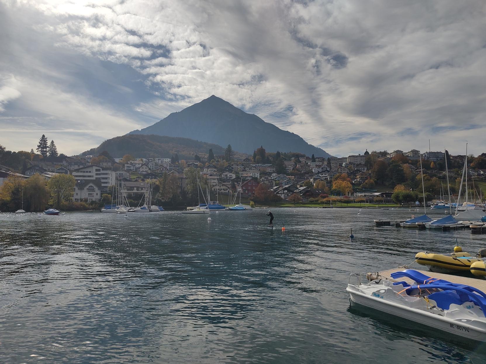 A dark pyramid-like silhouette looming over the harbour. Small boats and one foiler on the water. Trees in fall colours.