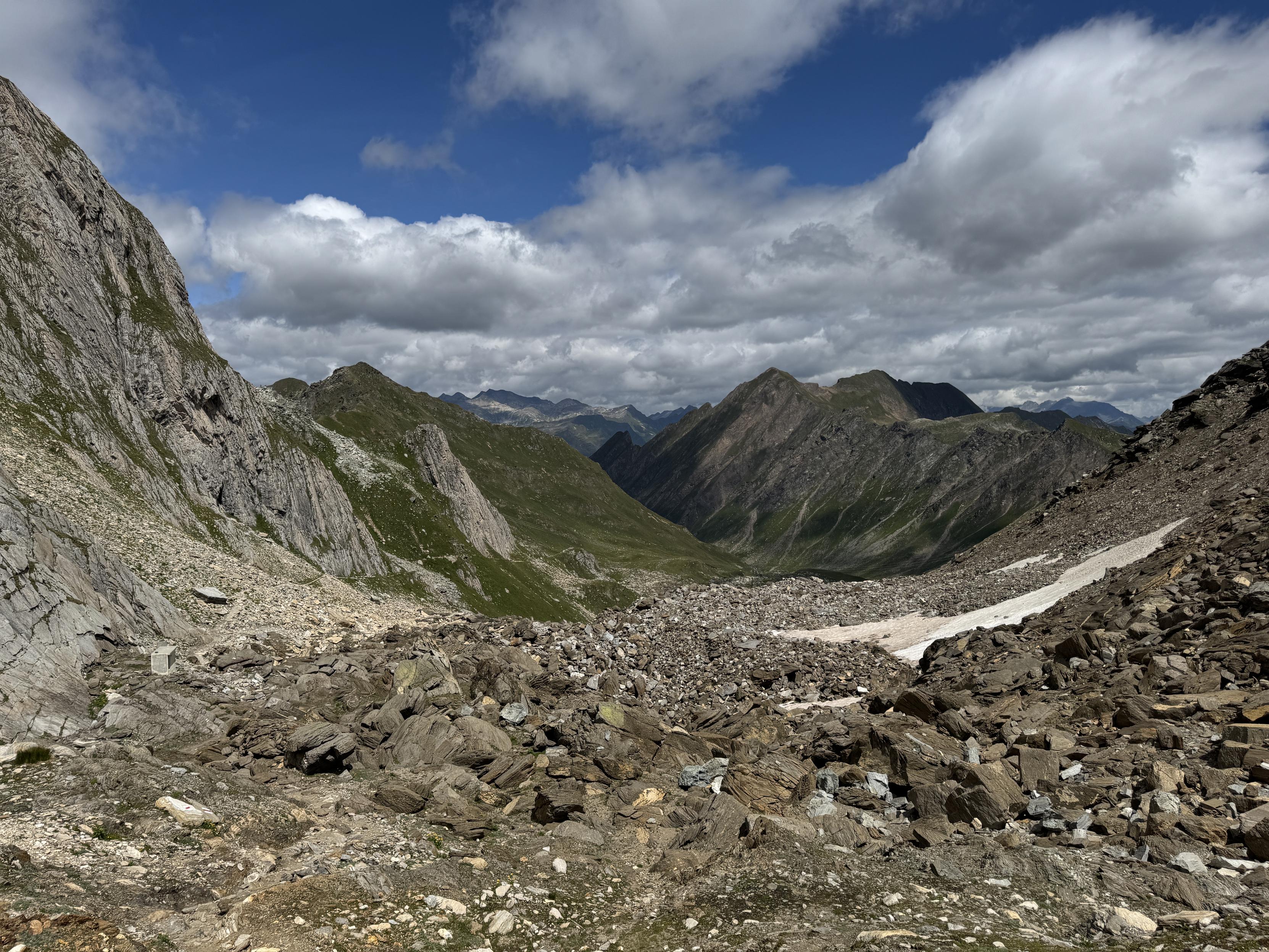 Blick von der Capanna Cristallina. Vorne recht steinig, in der Ferne felsige Berge mit etwas grün und ein weißblauer Himmel. 