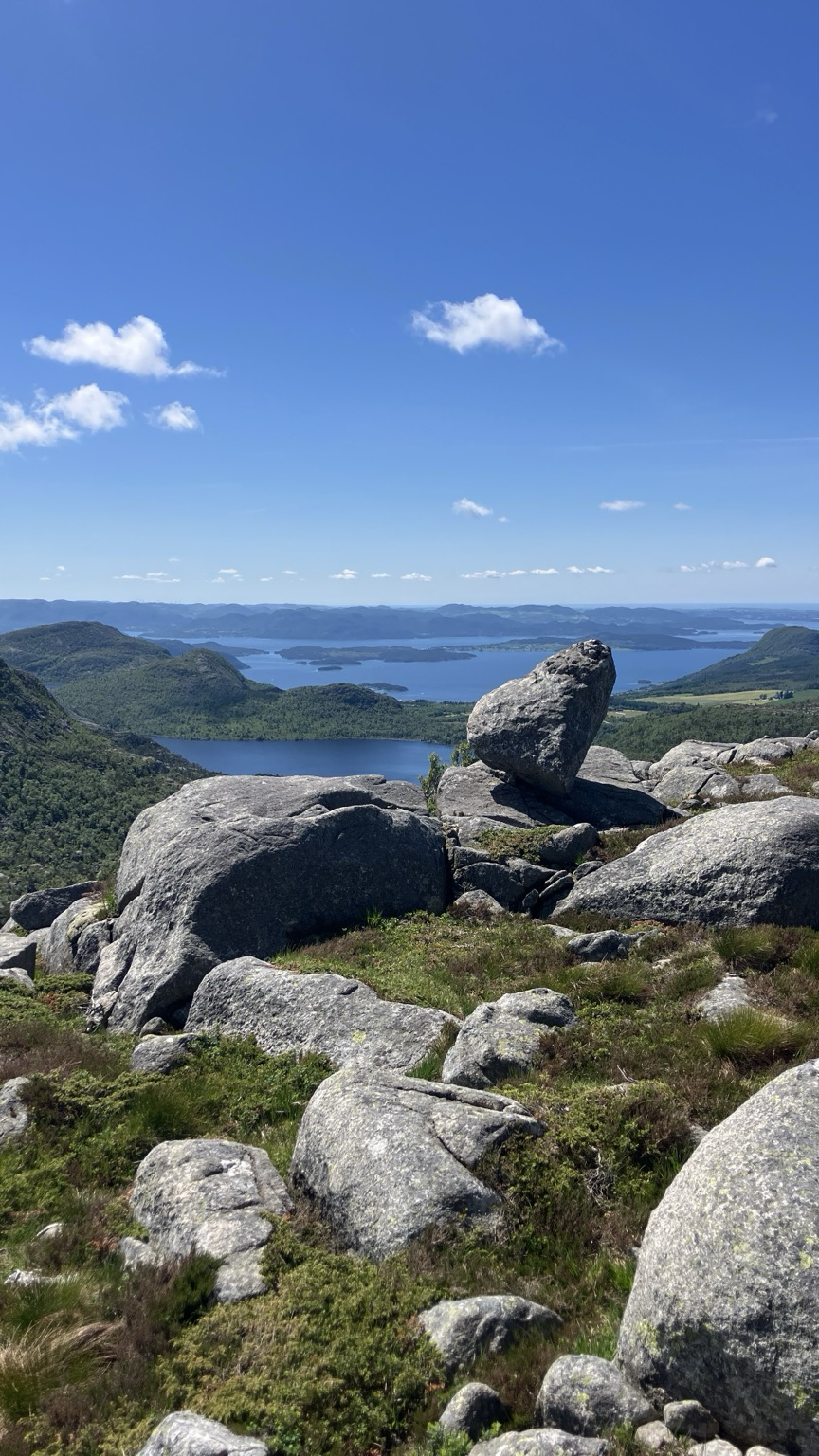 View from rocky terrain above Tysdalsvatnet in southern Norway, with a large boulder in the foreground and lakes, forested hills, and small islands in the distance under a clear blue sky.