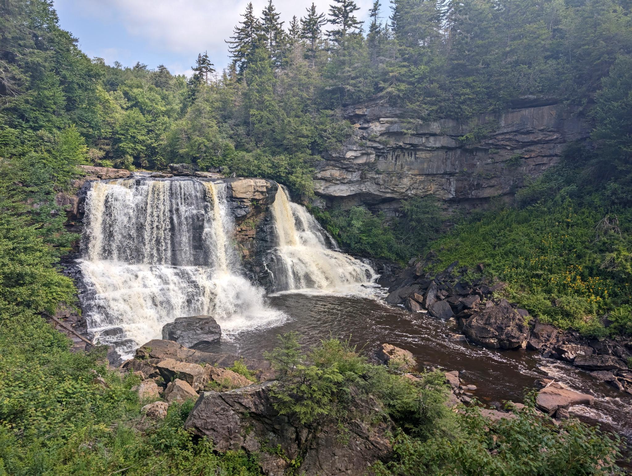 A wide, 57 foot waterfall cascading over layered rock cliffs into a pool below, surrounded by rocky cliffs and lush green trees and plants under a partly cloudy sky.