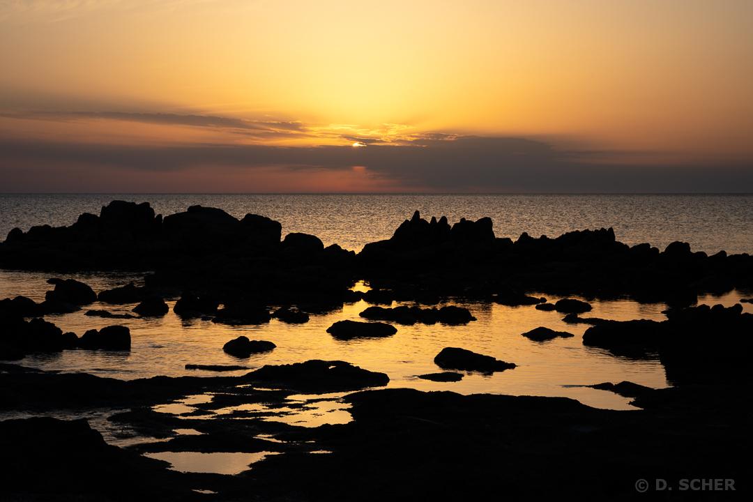 A string of silhouetted rocks separates the sea from a small stretch of water on the beach. The whole scene is bathed in the orange hues of the setting sun.
